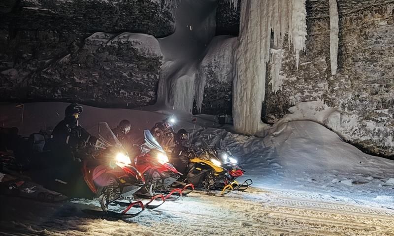 A line of snowmobiles are standing infront of a cliff with frozen ice hanging down