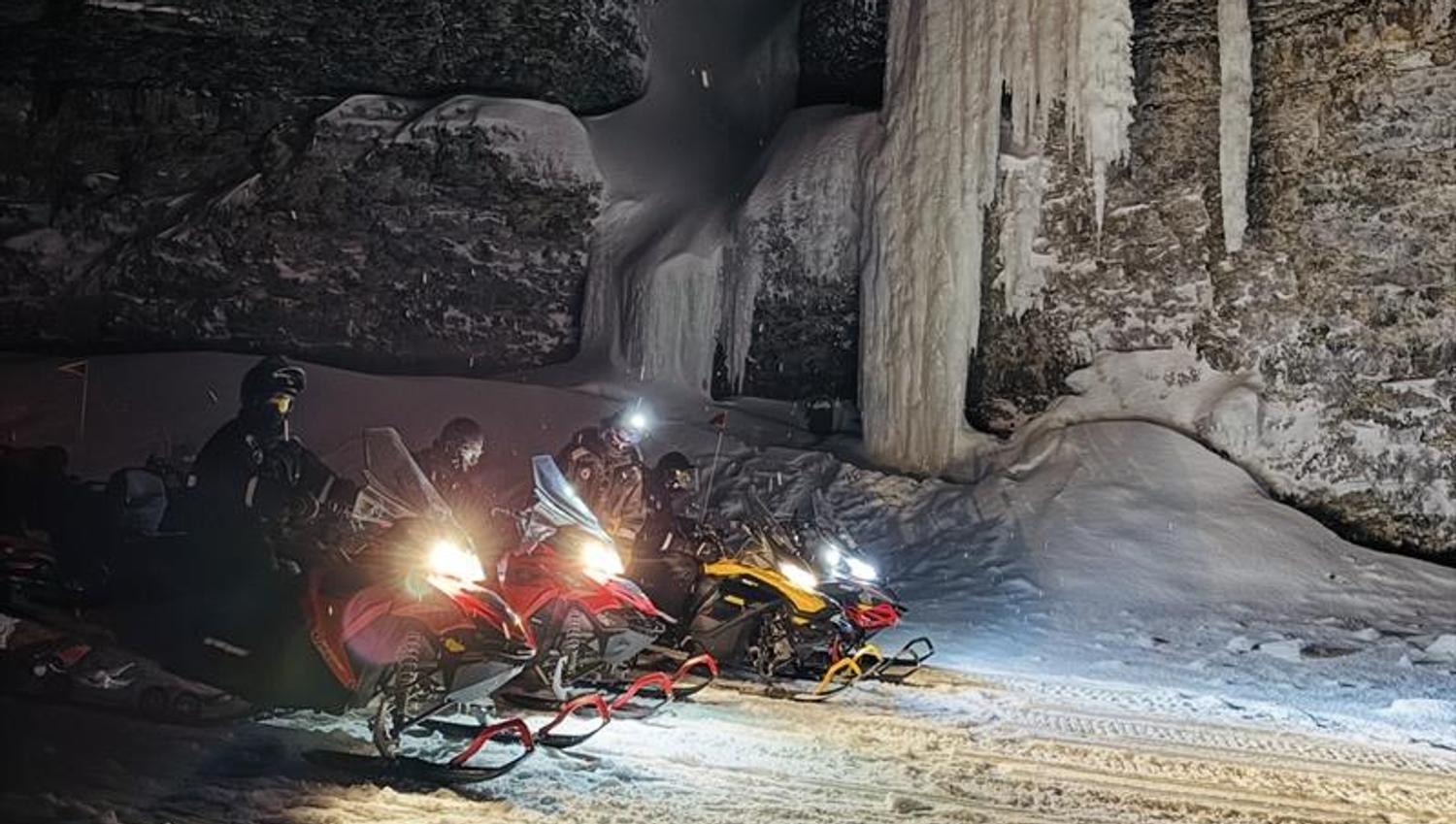 A line of snowmobiles are standing infront of a cliff with frozen ice hanging down