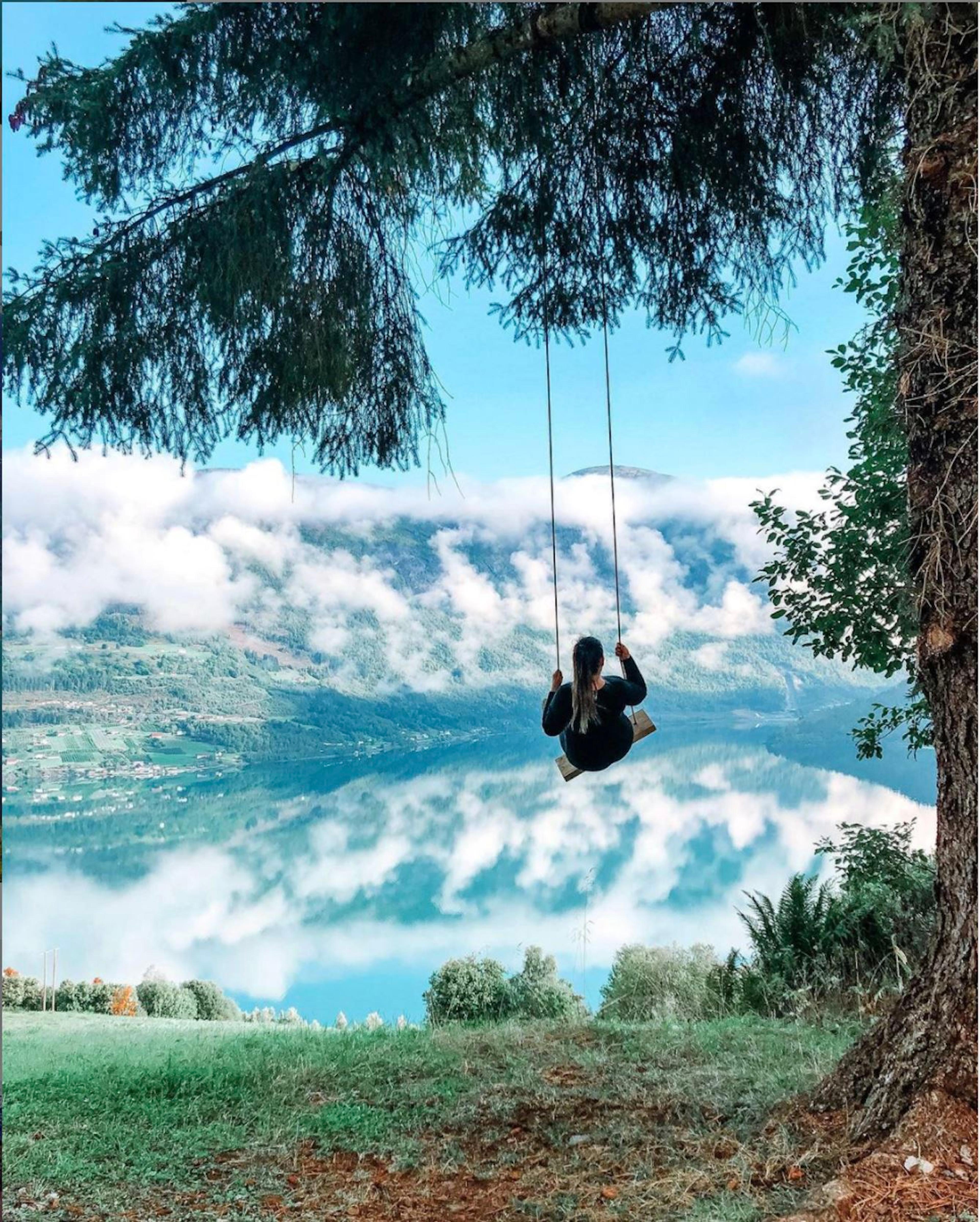 A person on a rope swing hangs from a tree above a grassy slope, overlooking a cloudy mountain lake at Skarsteinssætra i Olden.