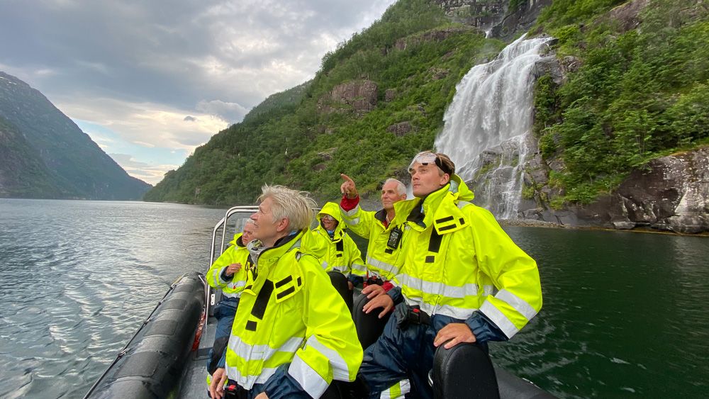 Turistar i gule drakter ser fascinert på eit imponerande fossefall frå RIB-båten i Hardangerfjorden.