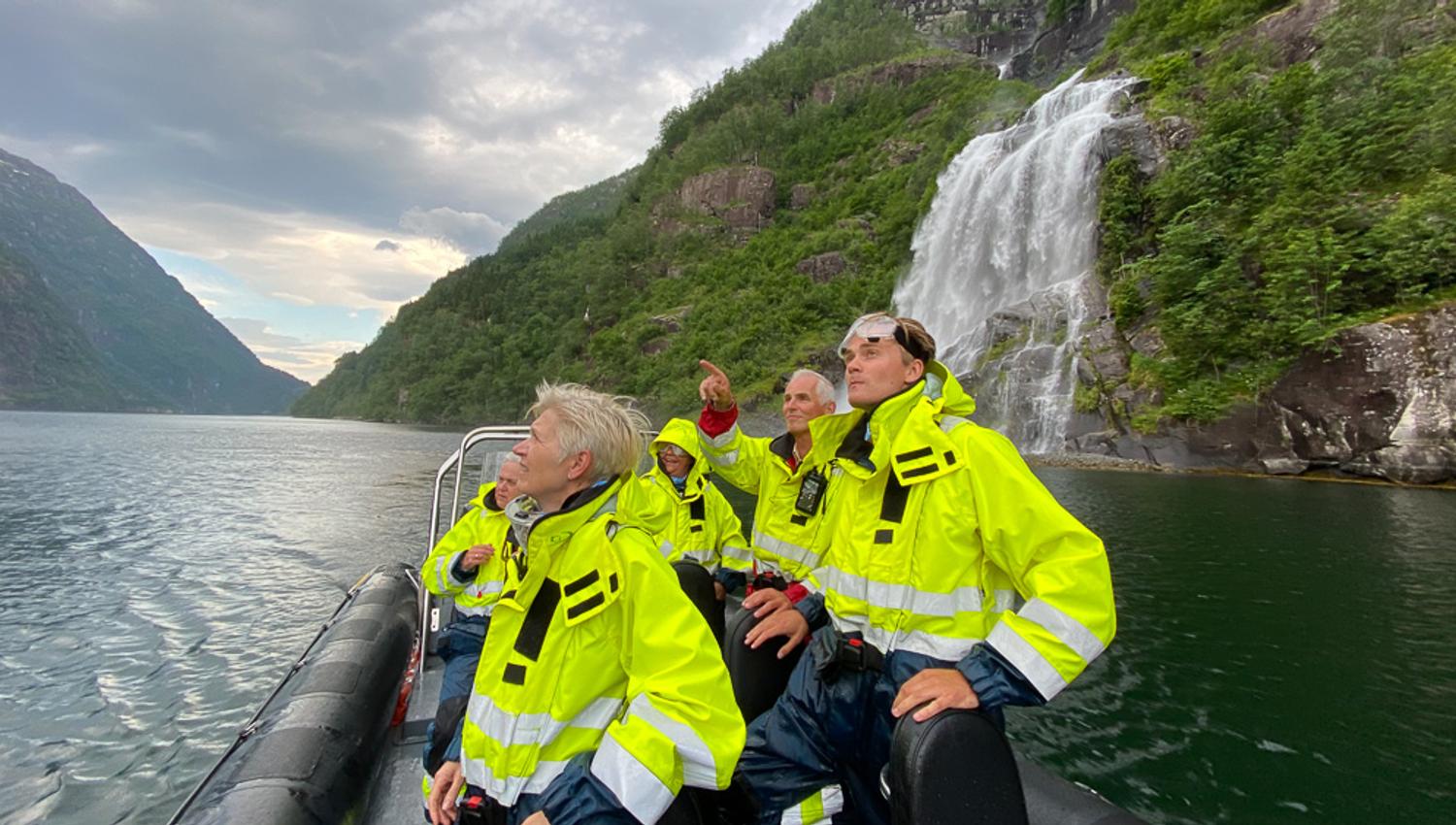 Turistar i gule drakter ser fascinert på eit imponerande fossefall frå RIB-båten i Hardangerfjorden.