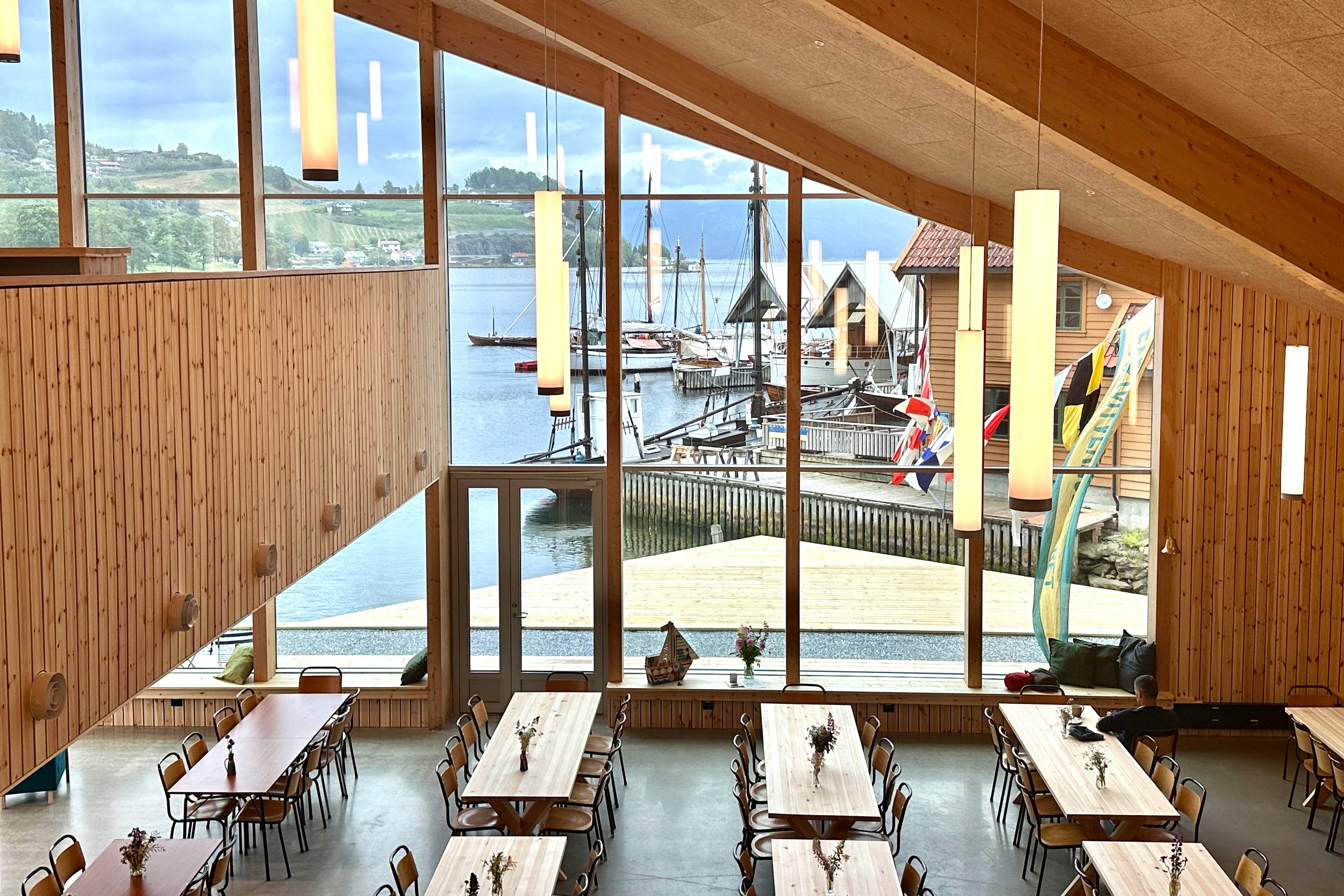 Large room with high ceilings and big windows overlooking the fjord. Many long tables are set up, and there is plenty of seating. View of the boat doc