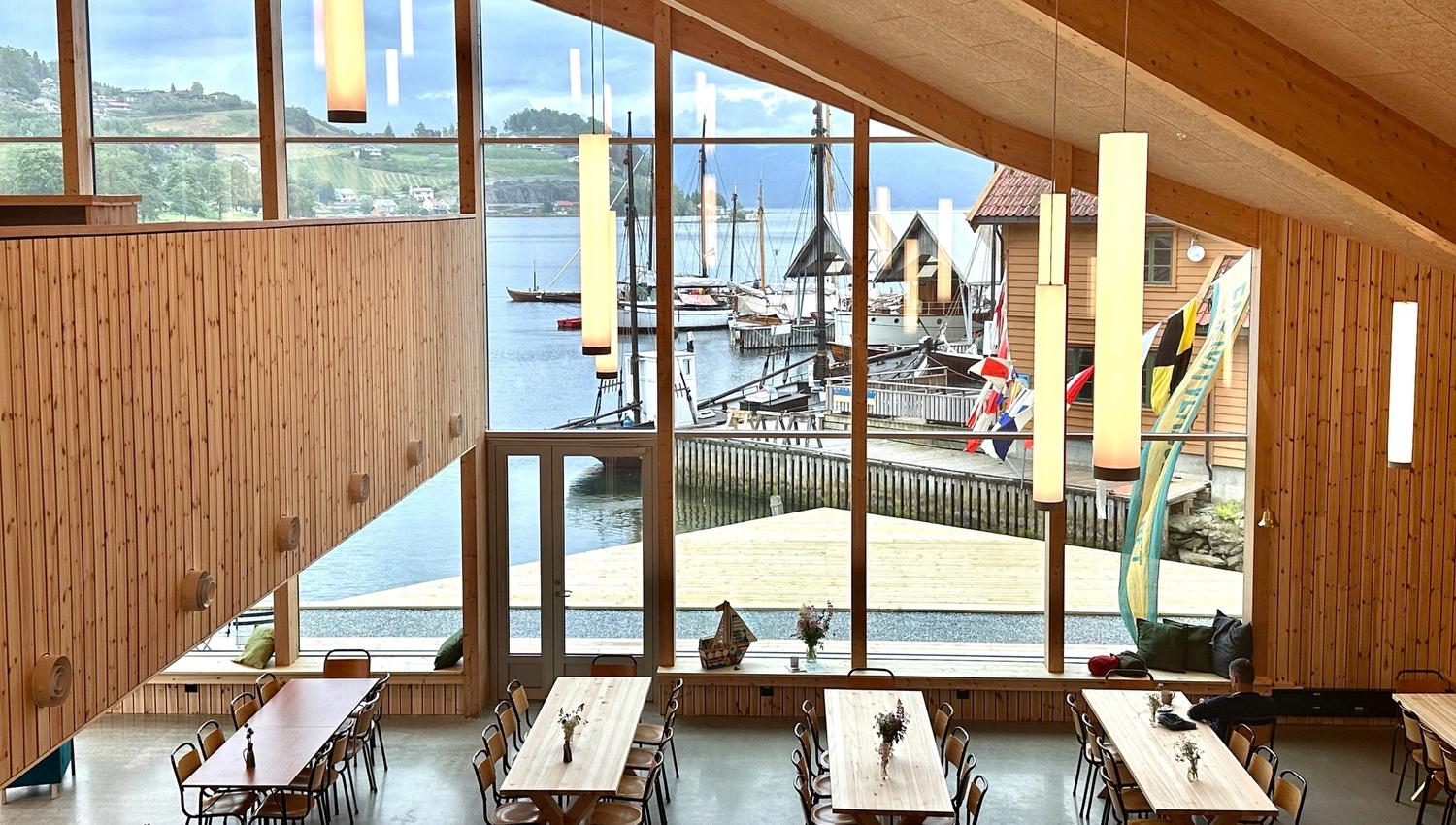 Large room with high ceilings and big windows overlooking the fjord. Many long tables are set up, and there is plenty of seating. View of the boat doc