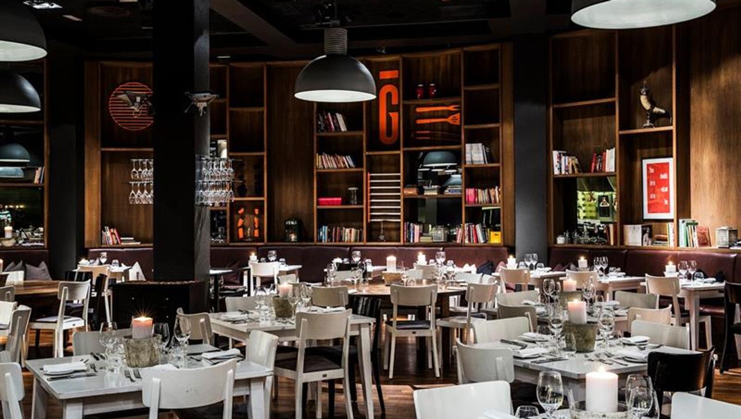 Restaurant interior: Small white tables and chairs, large bookshelves and gray ceiling lights.