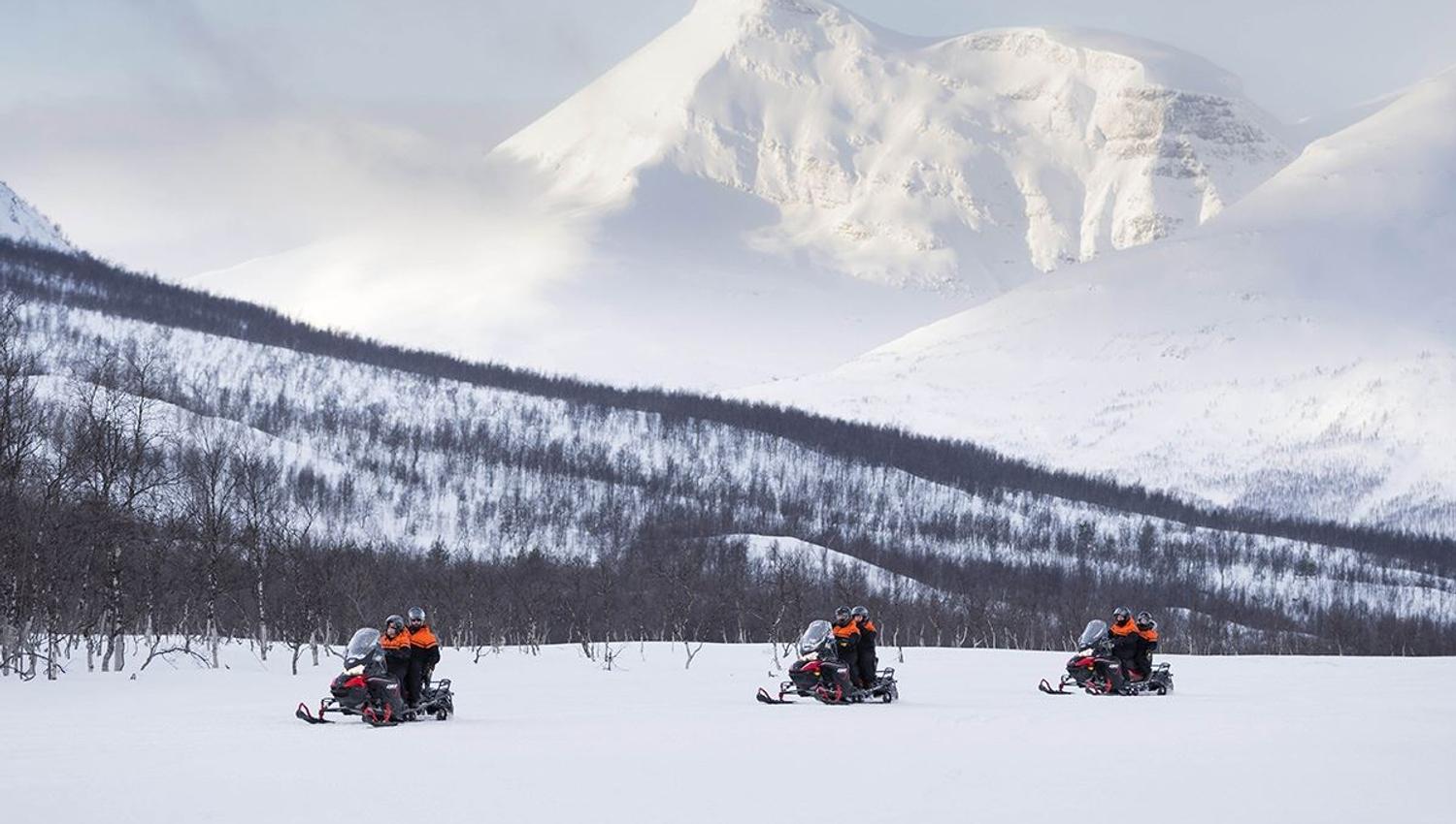 Three snow scooters drive past huge mountains