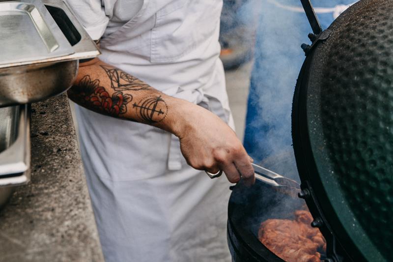 A cook wearing a hat with "breddos" written on the front is flipping meat on a round barbequeue.