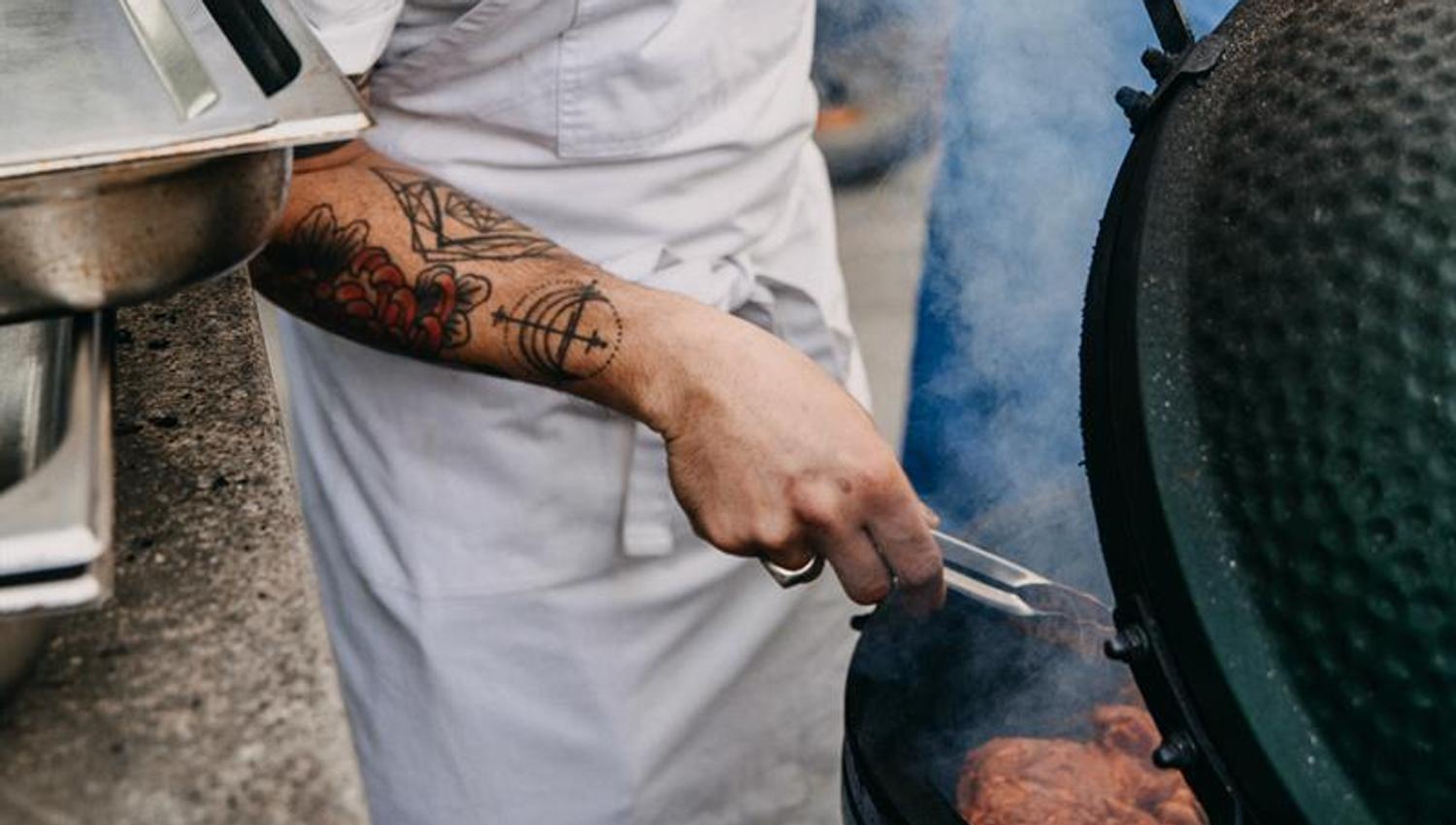A cook wearing a hat with "breddos" written on the front is flipping meat on a round barbequeue.