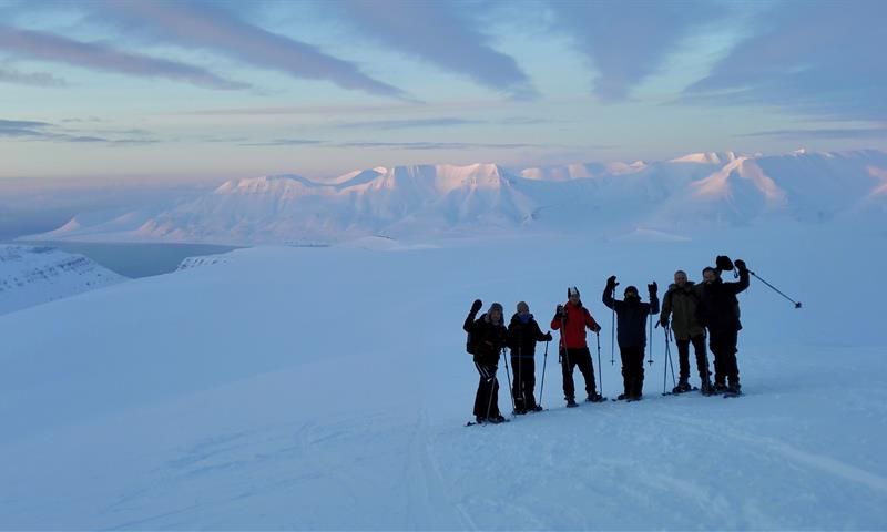 En gruppe med gjester på en snødekt fjellrygg i forgrunnen, med et vidstrakt snødekt landskap og fjerne fjell i bakgrunnen