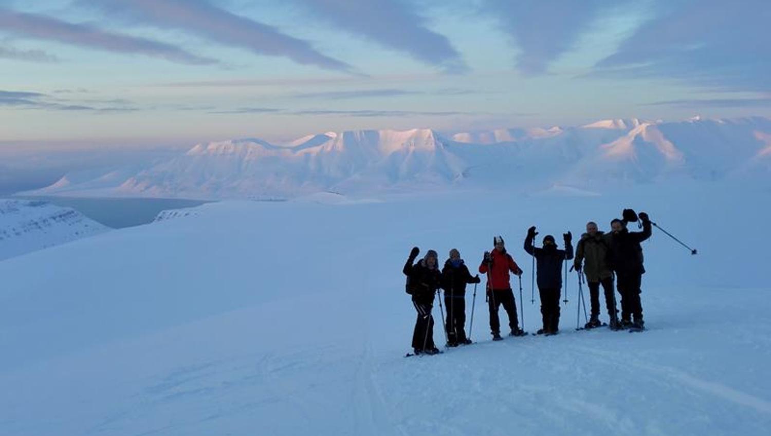 En gruppe med gjester på en snødekt fjellrygg i forgrunnen, med et vidstrakt snødekt landskap og fjerne fjell i bakgrunnen