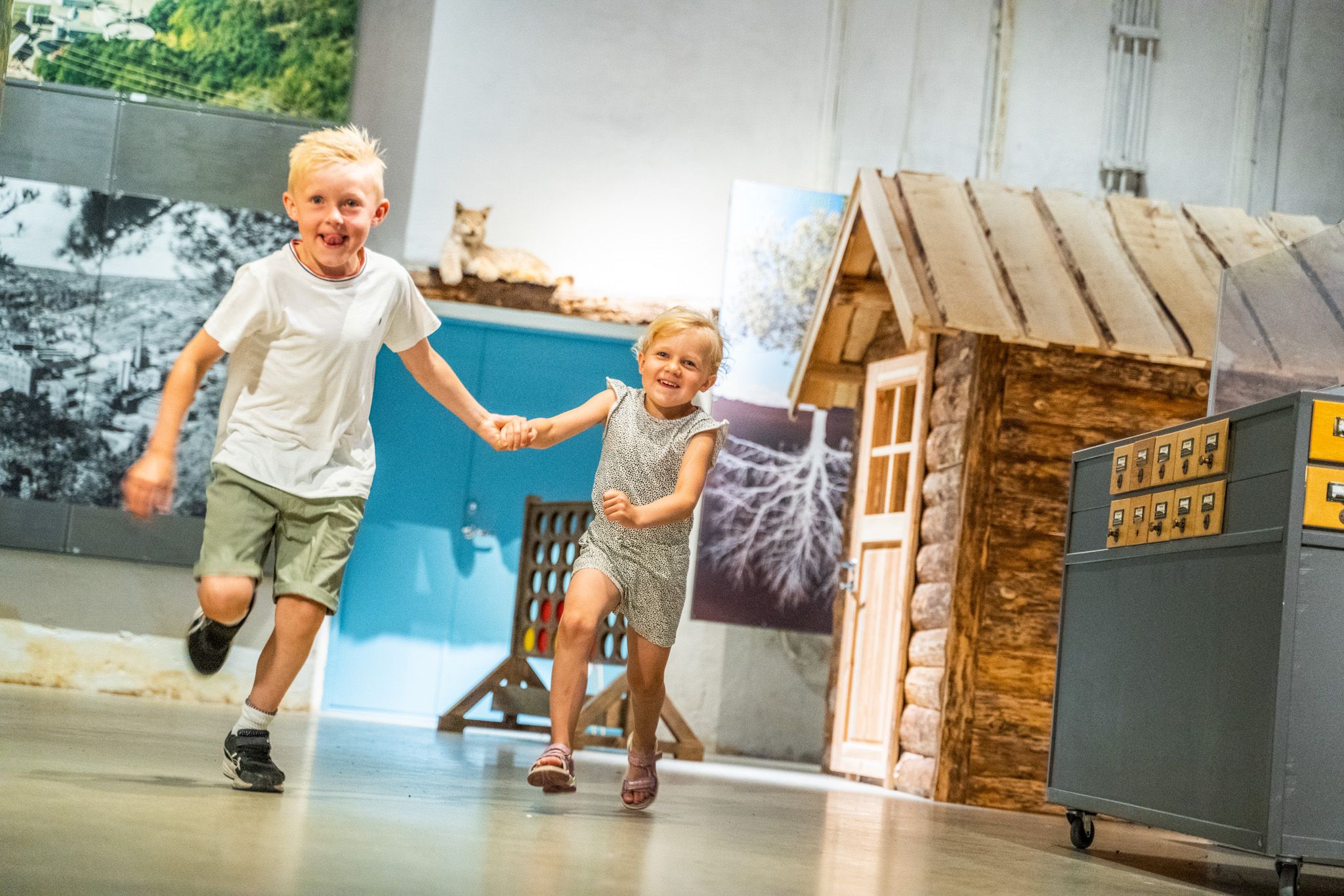 Bildet viser en gutt og ei jente som løper hånd i hånd og smiler. De ser glade ut og leker inne i et museum, der man kan se en liten tømmerhytte og noen utstillinger i bakgrunnen.