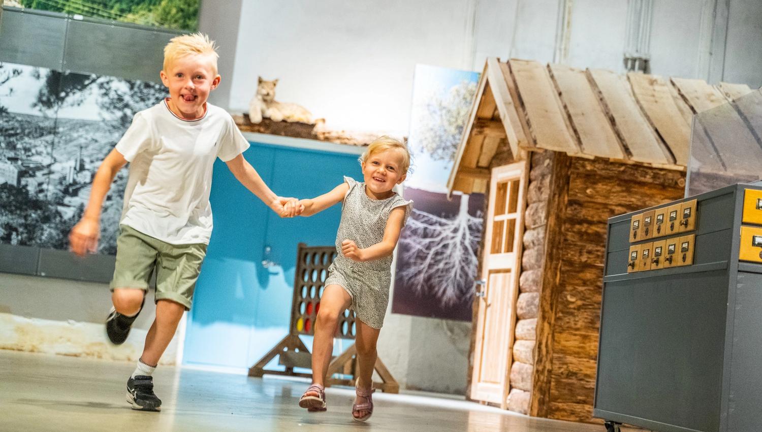 Bildet viser en gutt og ei jente som løper hånd i hånd og smiler. De ser glade ut og leker inne i et museum, der man kan se en liten tømmerhytte og noen utstillinger i bakgrunnen.