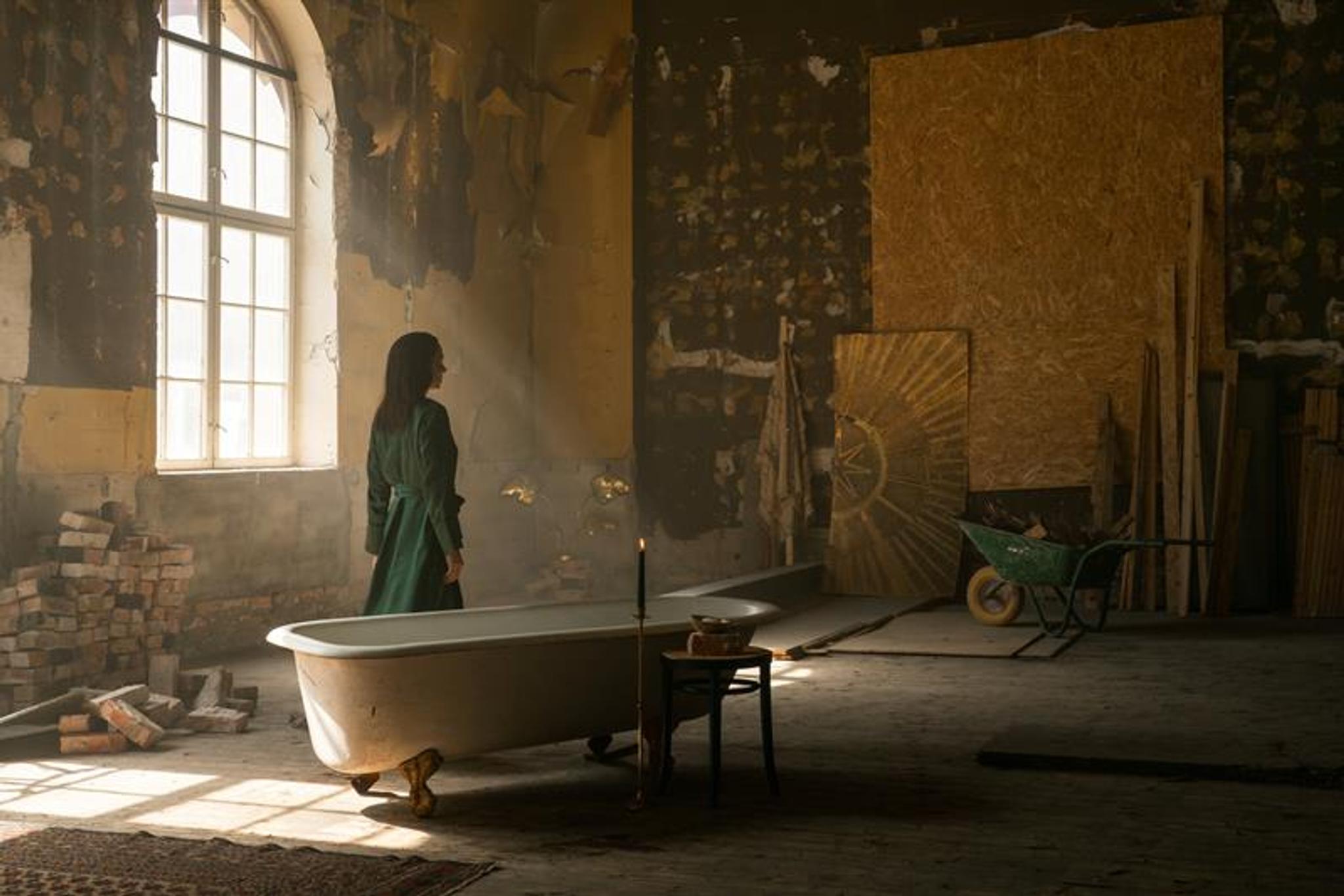 A woman in a green coat stands in a worn, dilapidated room beside a freestanding bathtub, surrounded by building materials and softly streaming light.