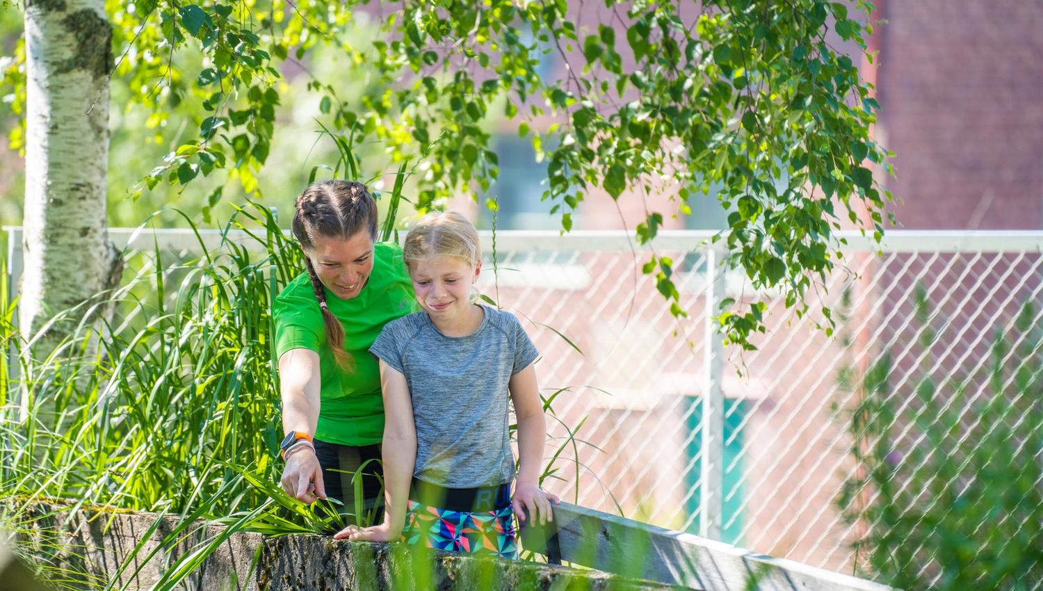 Bildet viser 2 jenter som står ute ved et gjerde omgitt av grønne planter. Ene jenta peker på noe i vannet eller gresset, og jenta ser nysgjerrig dit hun peker. Det er sol og trær rundt dem.