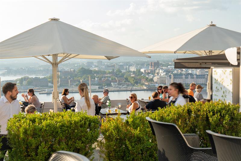 Guests enjoy food and drinks on a sunny rooftop terrace with panoramic views over Oslo and the fjord.