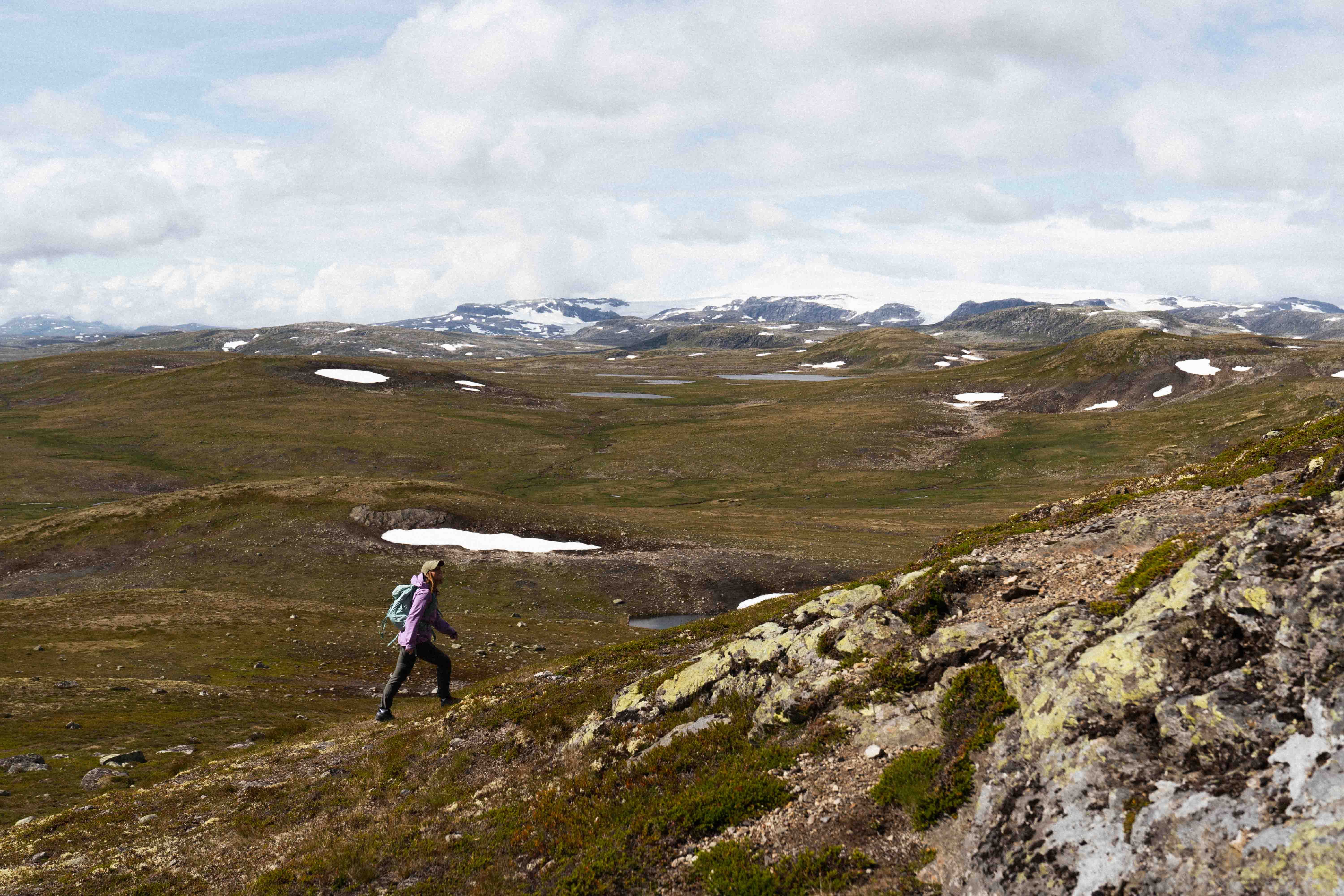 A female hiker hiking uphill towards the Halnekollen summit, the Hardangerjøkule glacier is seen in the background.