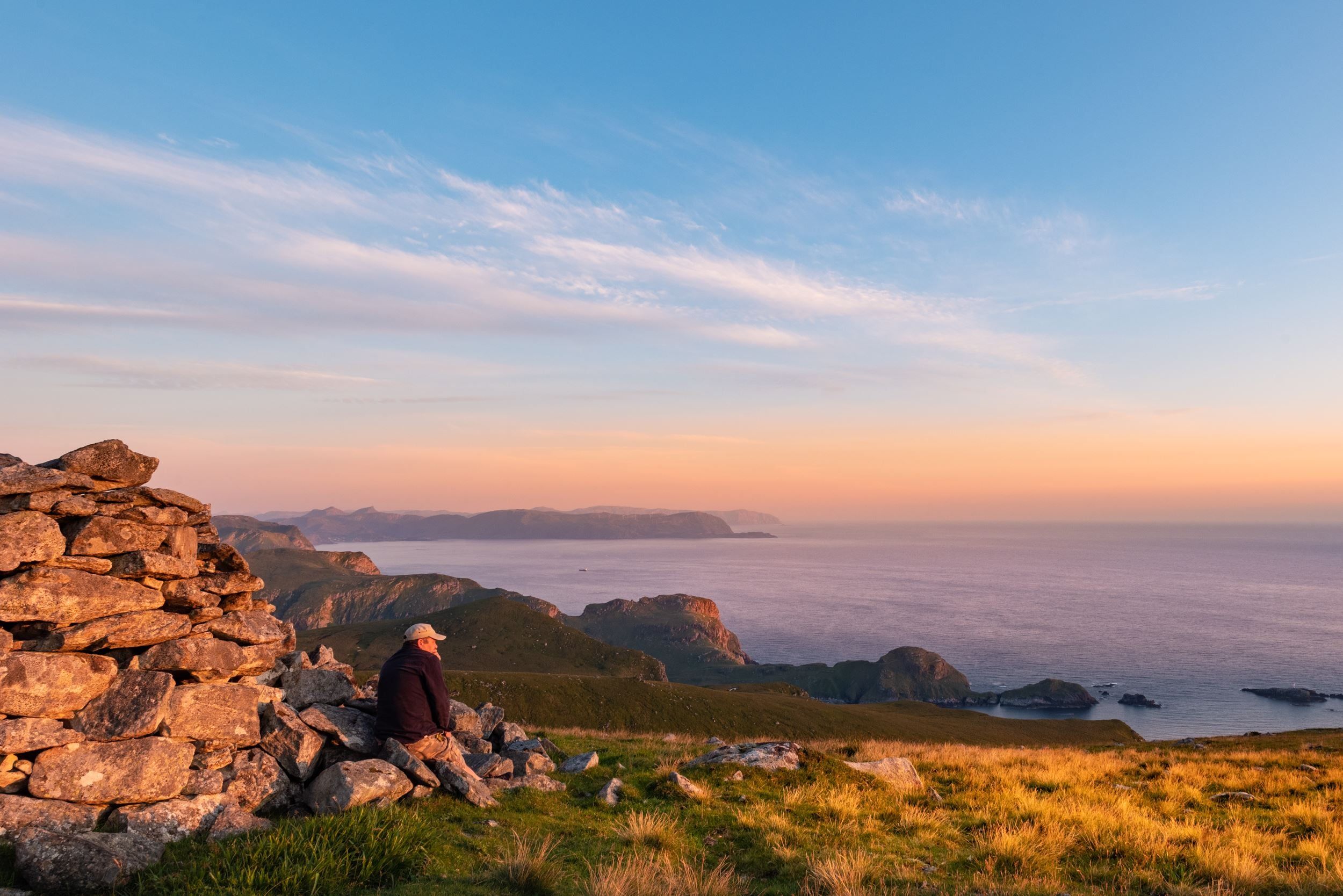 Ein person sit ved ein steinvarde på ei grasdekt høgd og ser utover havet og øyane i kveldssol..