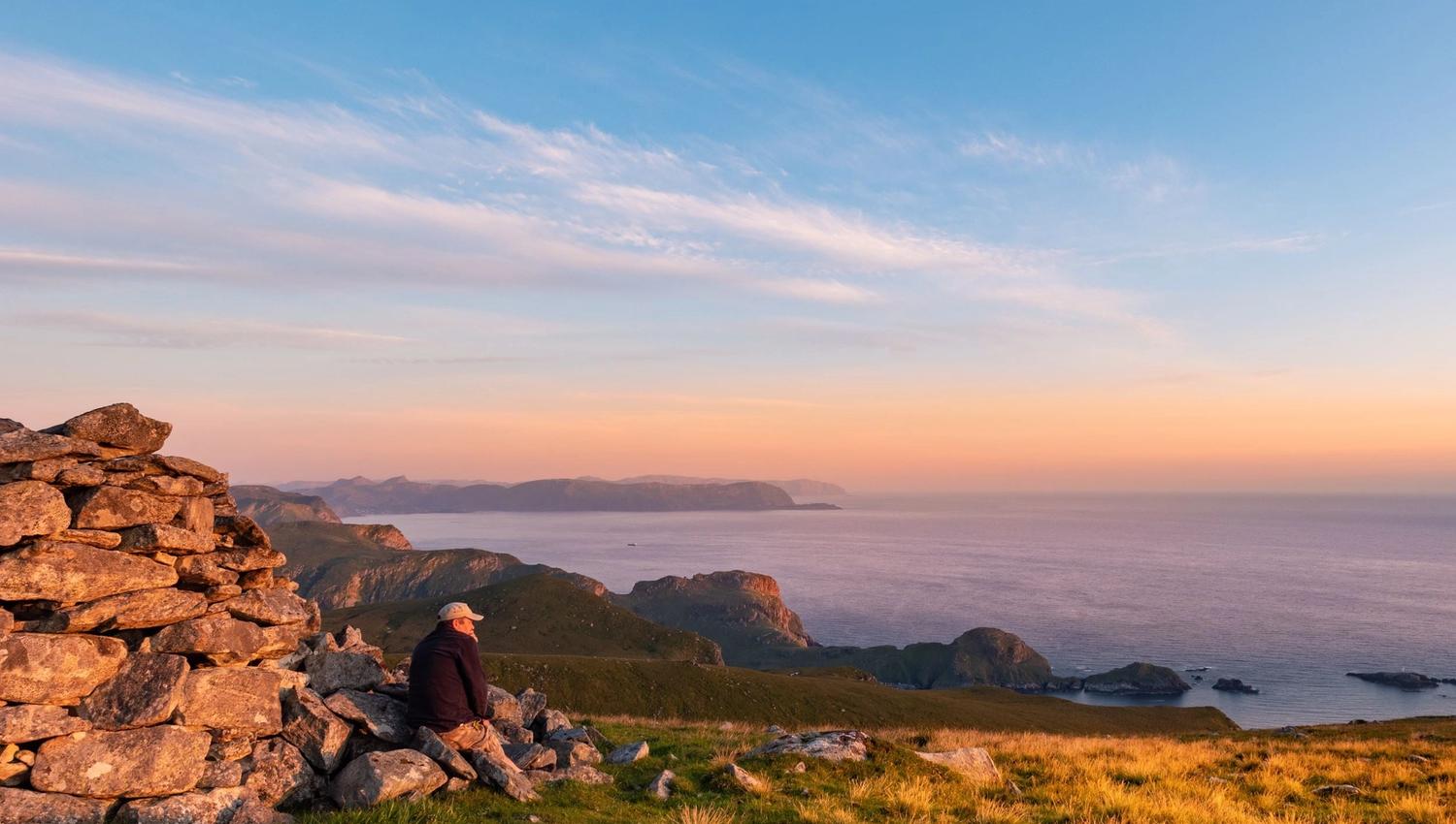 Ein person sit ved ein steinvarde på ei grasdekt høgd og ser utover havet og øyane i kveldssol..