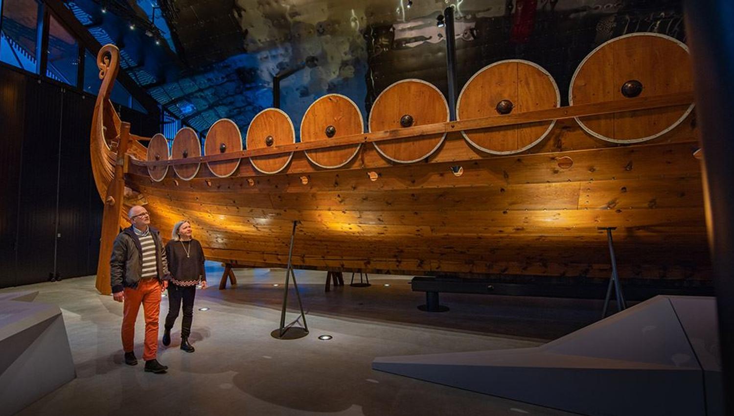 Stort vikingskip i tre med runde skjold langs rekka, utstilt innandørs i eit museum, Sagastad, medan to besøkjande går forbi..