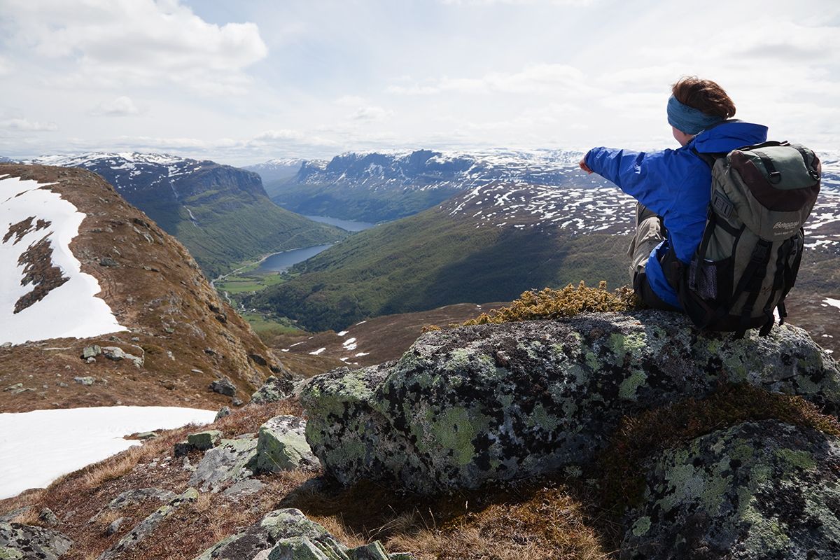 A woman sits on a rock and enjoyes a magnificent view into a steep valley with a lake on the bottom.