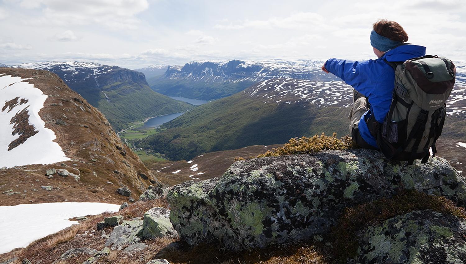 A woman sits on a rock and enjoyes a magnificent view into a steep valley with a lake on the bottom.