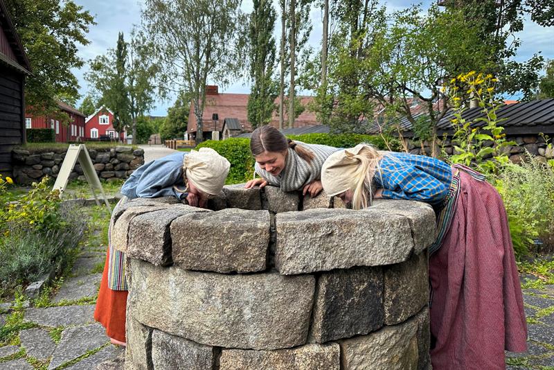 tre jenter som kikker ned i en brønn, de er kledde i gammeldagse klær