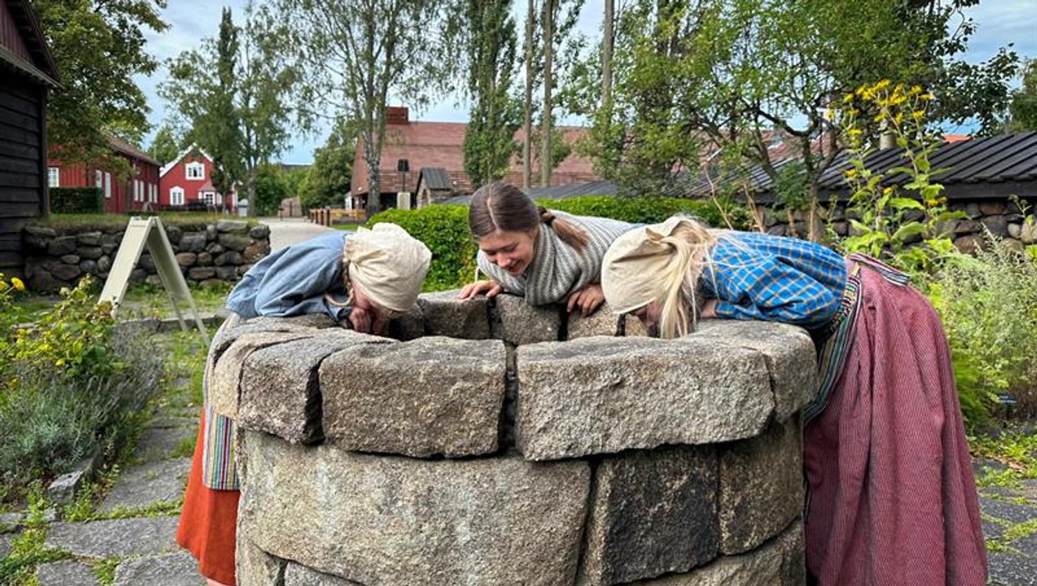 tre jenter som kikker ned i en brønn, de er kledde i gammeldagse klær