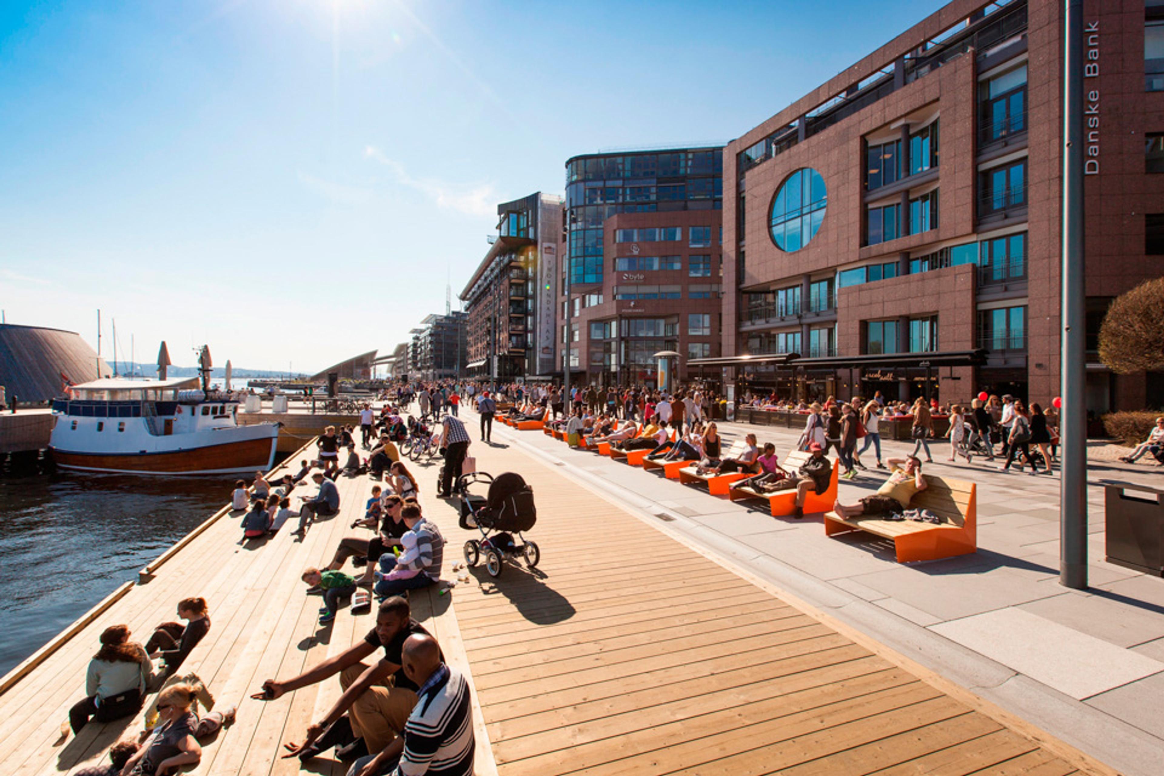 Sunny, blue sky, people sitting alongside the pier, a boat on the left hand side, brown buildings with large windows on the right hand side. People wa