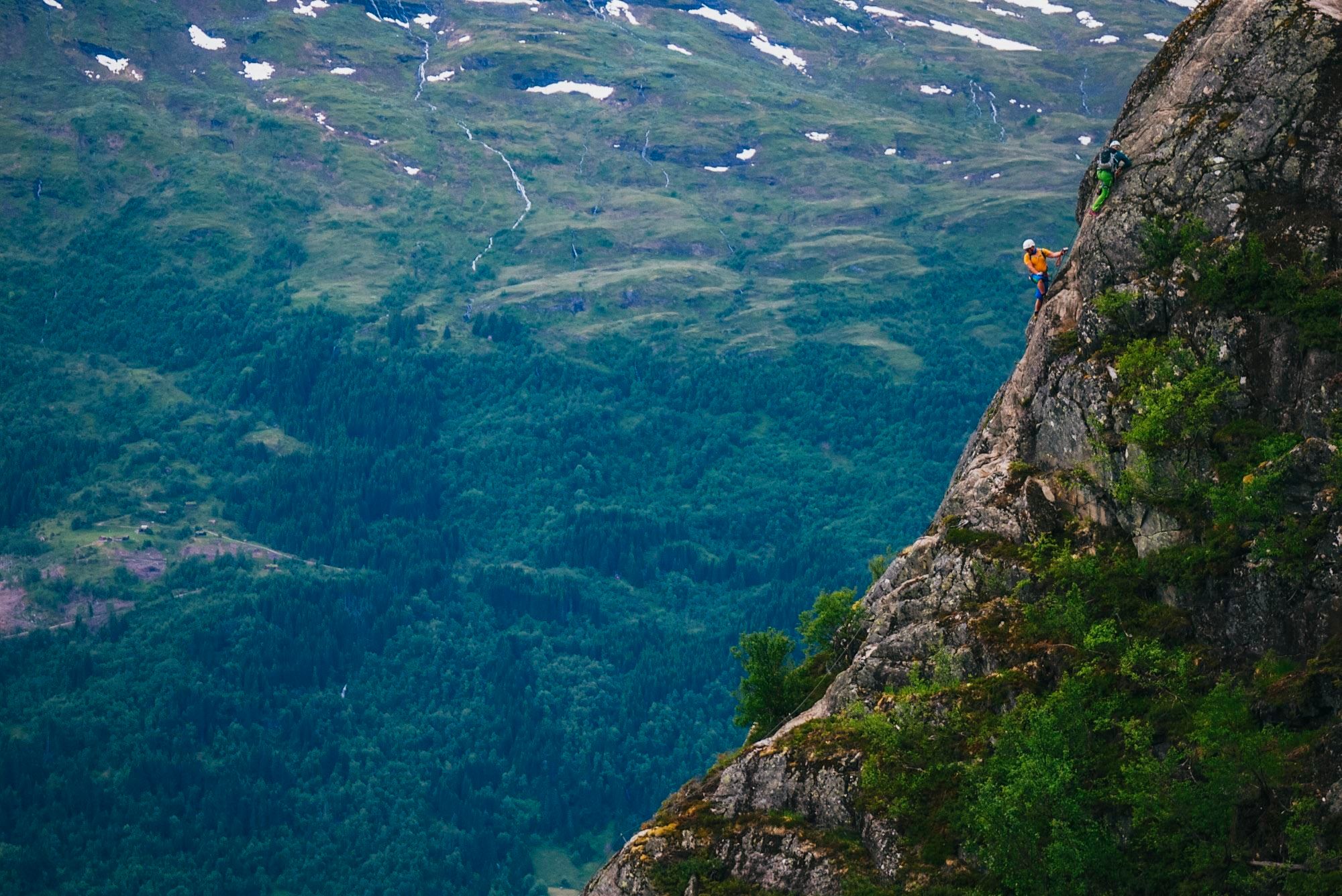 To klatrarar med hjelm på ei bratt fjellside over grøne skogar og fjellandskap med snøflekkar..