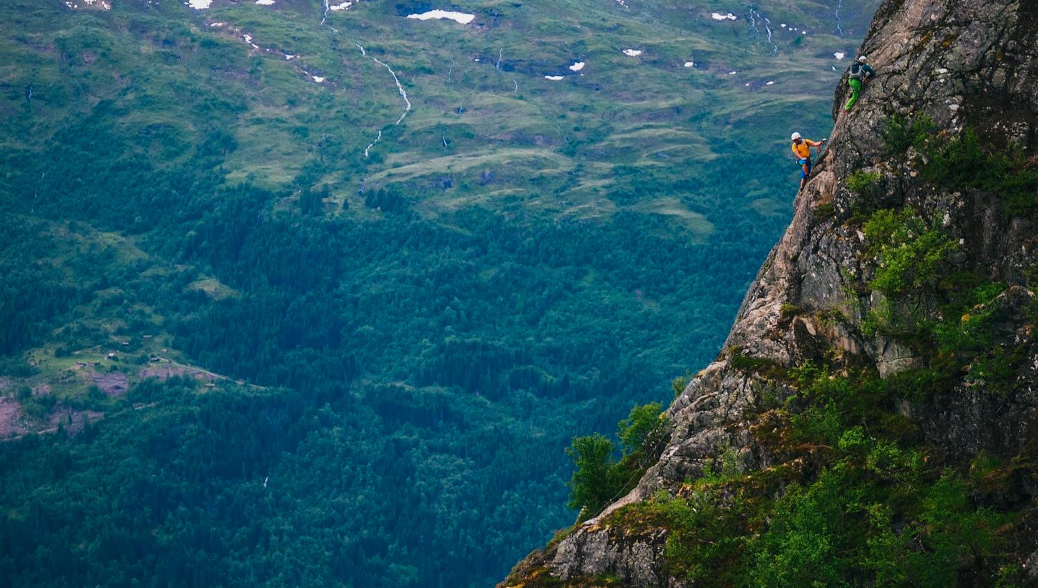 To klatrarar med hjelm på ei bratt fjellside over grøne skogar og fjellandskap med snøflekkar..
