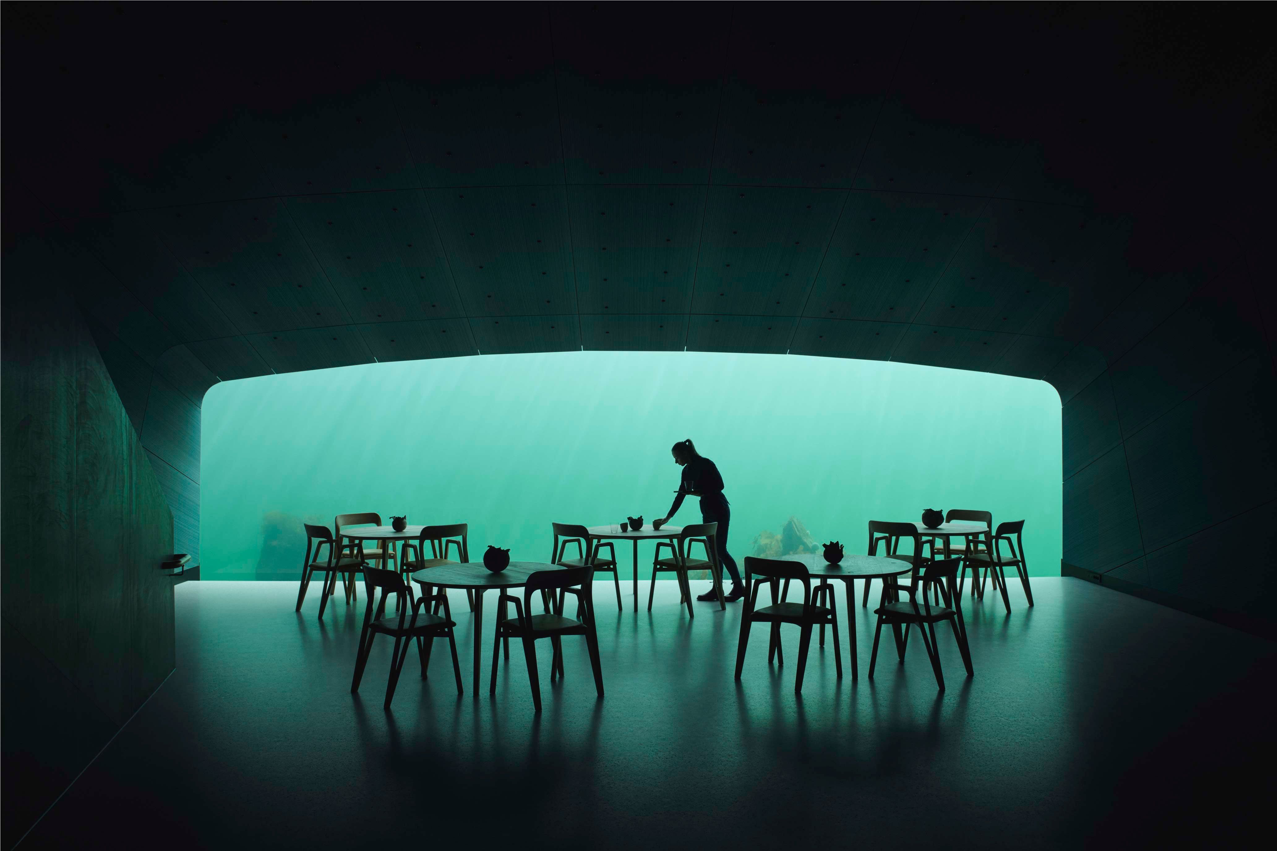 An employee is setting the tables, a dim room with turquoise light from the sea flowing into the dining area.

