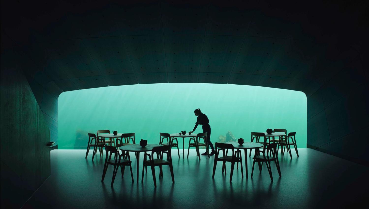 An employee is setting the tables, a dim room with turquoise light from the sea flowing into the dining area.