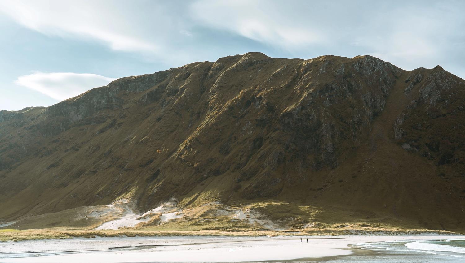 Stor fjellrygg reiser seg over ei lys sandstrand i Ervik med grunt vatn, under lettskyet himmel i mjukt lys..