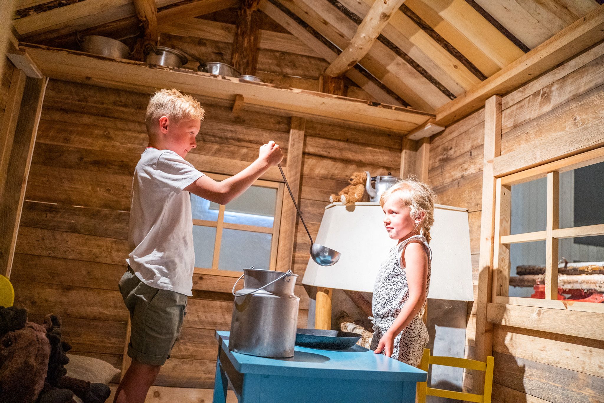 Bildet viser en gutt og ei jente som leker inne i et lite trehus. Gutten holder en øse over en stor melkekanne, mens jenta ser på og smiler. Det ser ut som de later som de lager mat eller heller melk. Stemningen er lys og koselig.