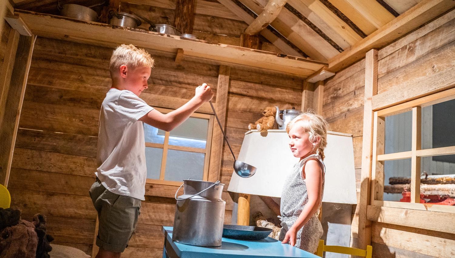 Bildet viser en gutt og ei jente som leker inne i et lite trehus. Gutten holder en øse over en stor melkekanne, mens jenta ser på og smiler. Det ser ut som de later som de lager mat eller heller melk. Stemningen er lys og koselig.