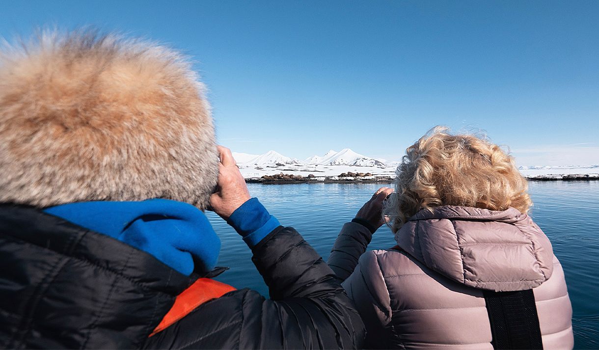 Two guests in the foreground standing on a boat while observing walruses on land in the background from a safe distance using binoculars