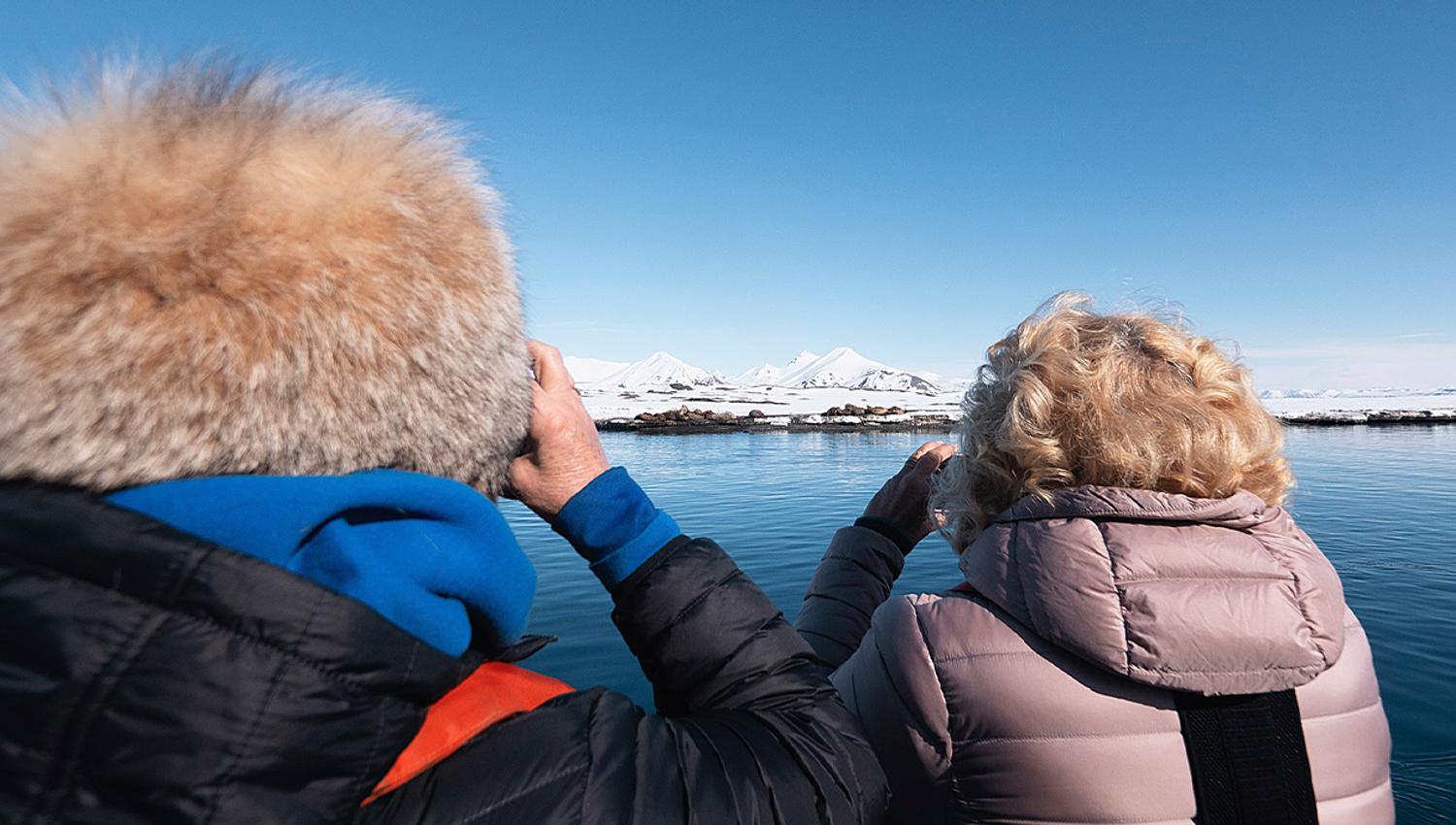 Two guests in the foreground standing on a boat while observing walruses on land in the background from a safe distance using binoculars