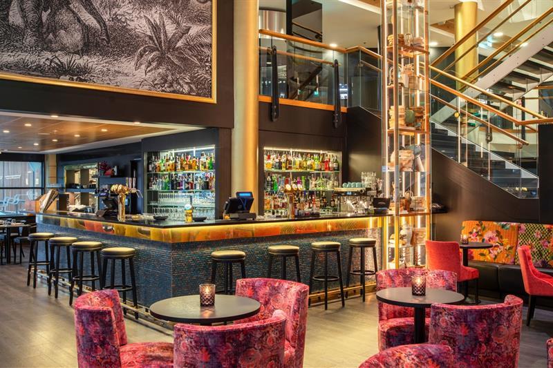 Bar med barstoler, små runde bord med røde stoler og trapp i bakgrunnen