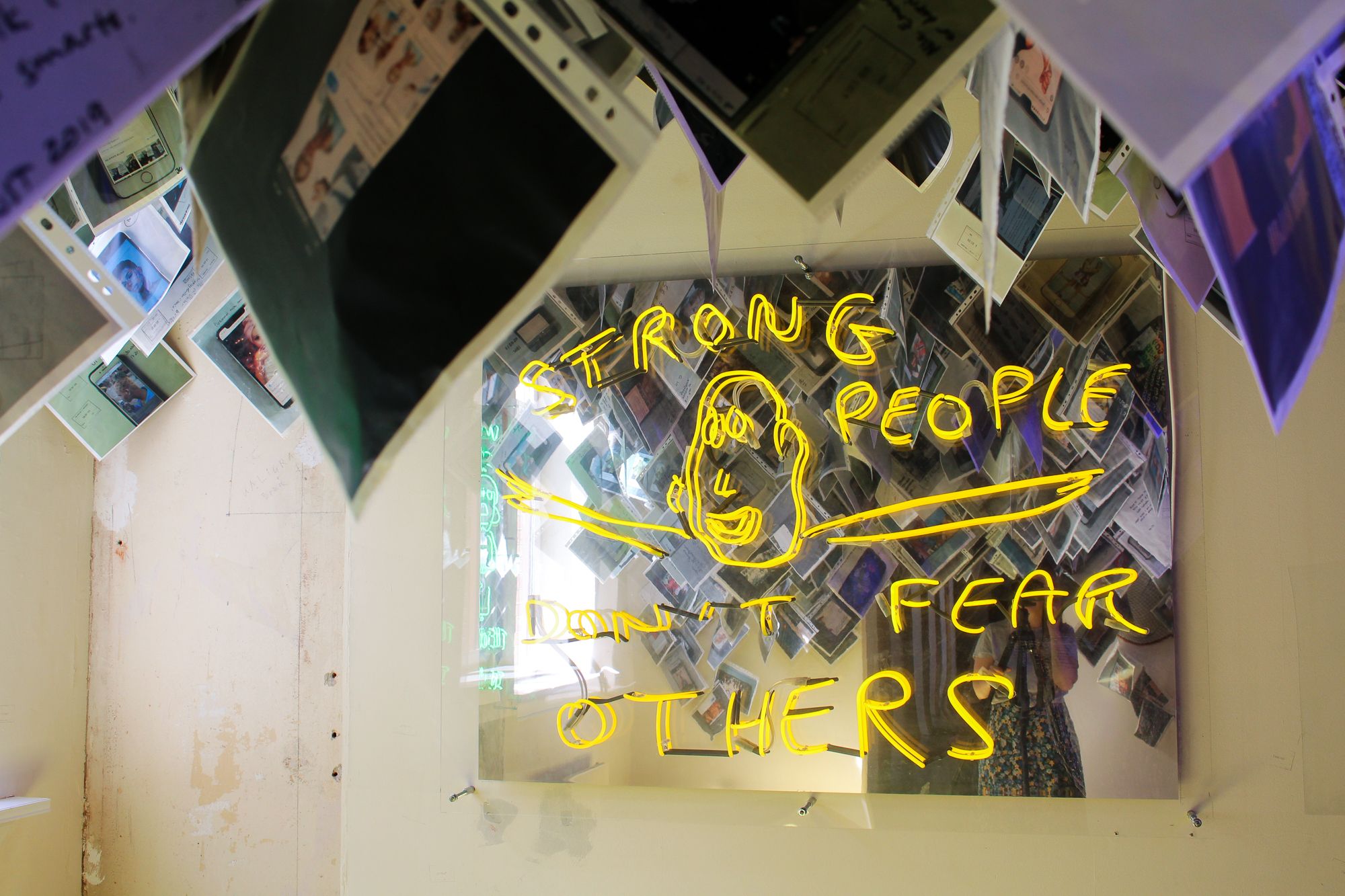 Diverse fotografier som henger fra taket, et neonskilt hvor det står "Strong people don't fear others".