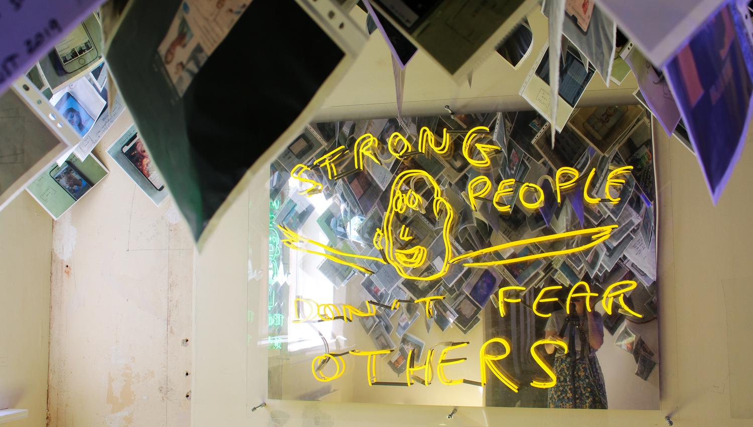 Diverse fotografier som henger fra taket, et neonskilt hvor det står "Strong people don't fear others".