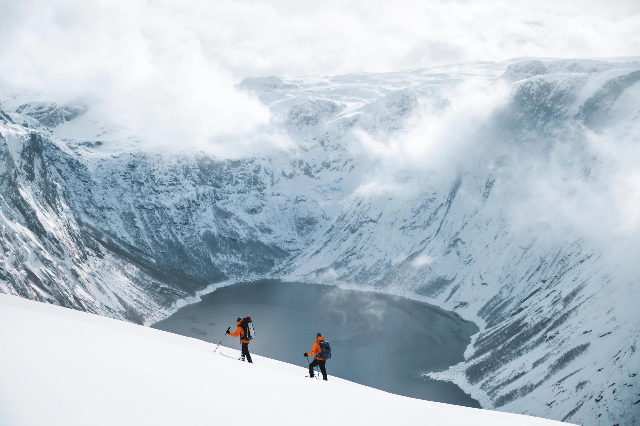 Two people skiing beside a lake surrounded by snowy mountains.