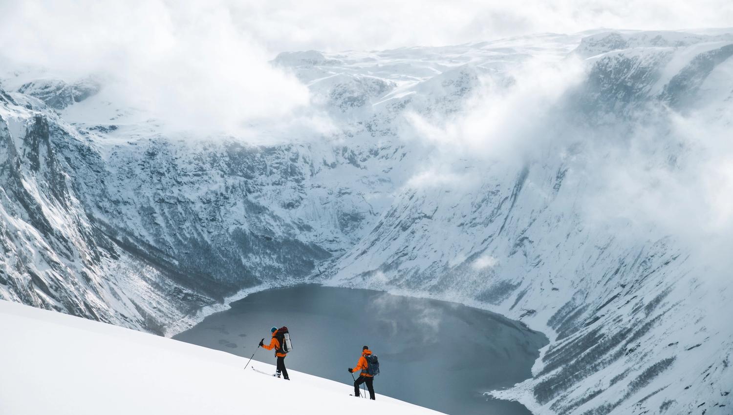 To skigåere ved et vann omringet av snøkledde fjell.