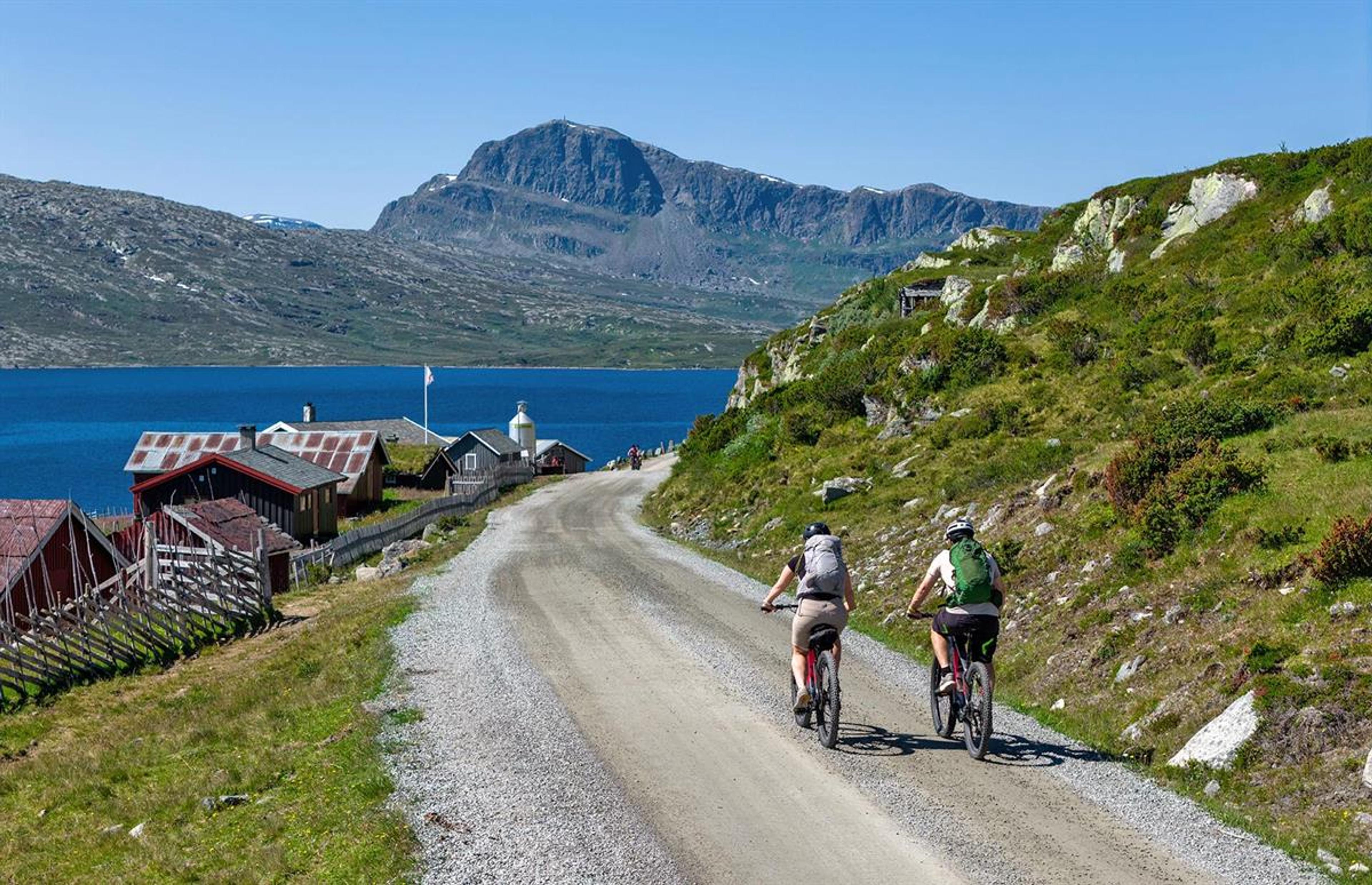 To syklister på grusvei langs vann med fjell i bakgrunnen.