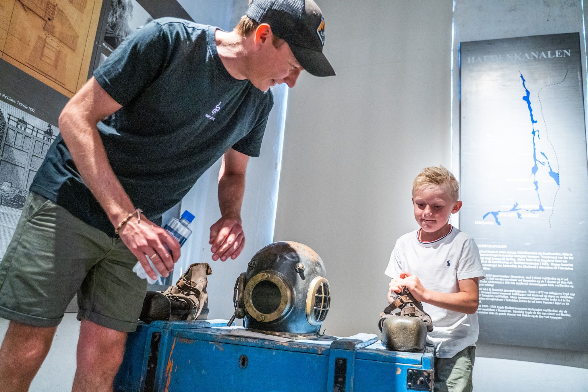 Bildet viser en mann og en gutt som ser på gamle dykkerutstyr i et museum. På bordet foran dem ligger en tung, gammeldags dykkerhjelm og sko av metall. Gutten smiler mens han ser på utstyret, og mannen forklarer noe for ham.