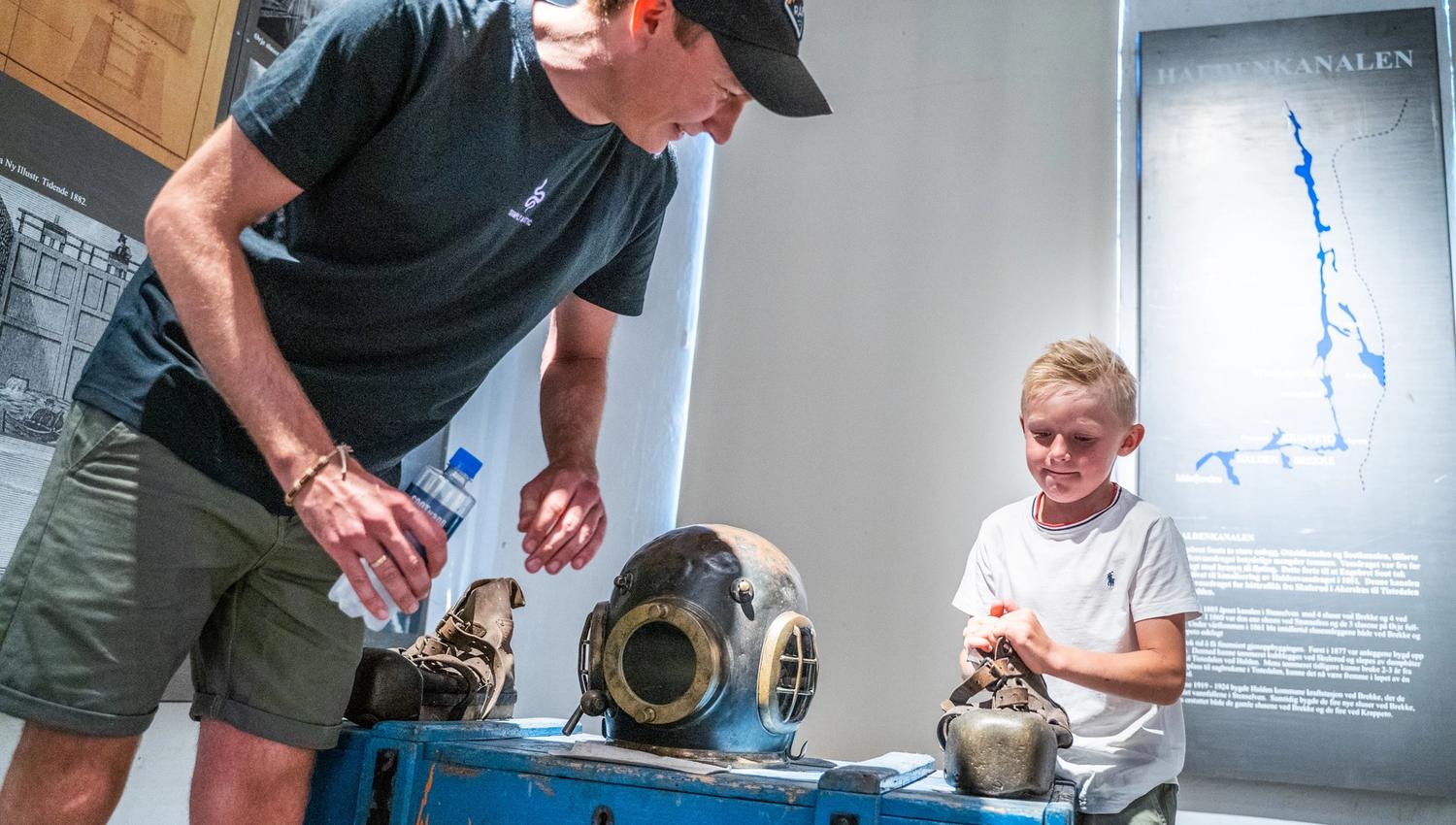Bildet viser en mann og en gutt som ser på gamle dykkerutstyr i et museum. På bordet foran dem ligger en tung, gammeldags dykkerhjelm og sko av metall. Gutten smiler mens han ser på utstyret, og mannen forklarer noe for ham.