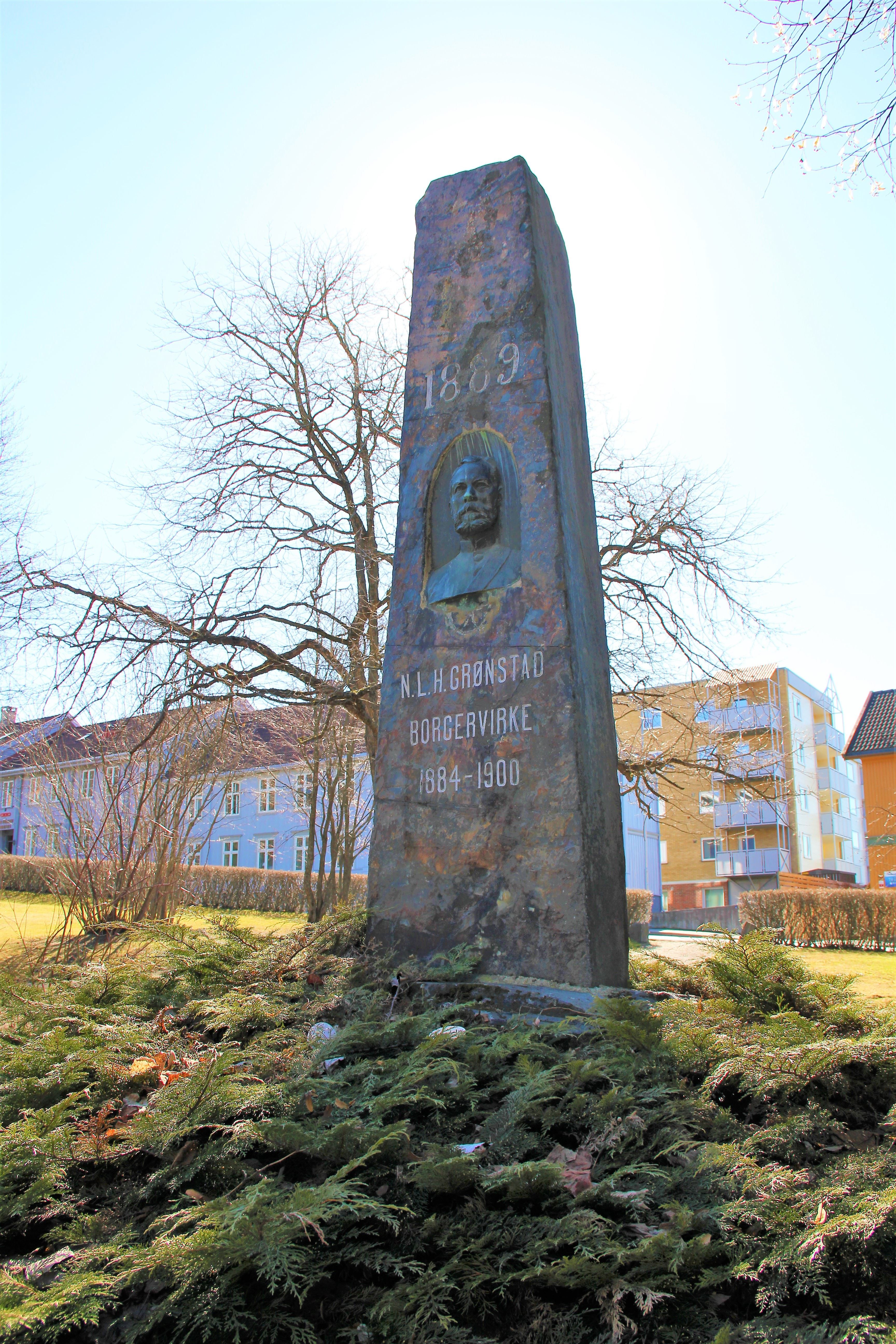 Høy steinmonument med inngravert tekst og nisje, omgitt av lave busker og trær ved boligblokker i sollys..
