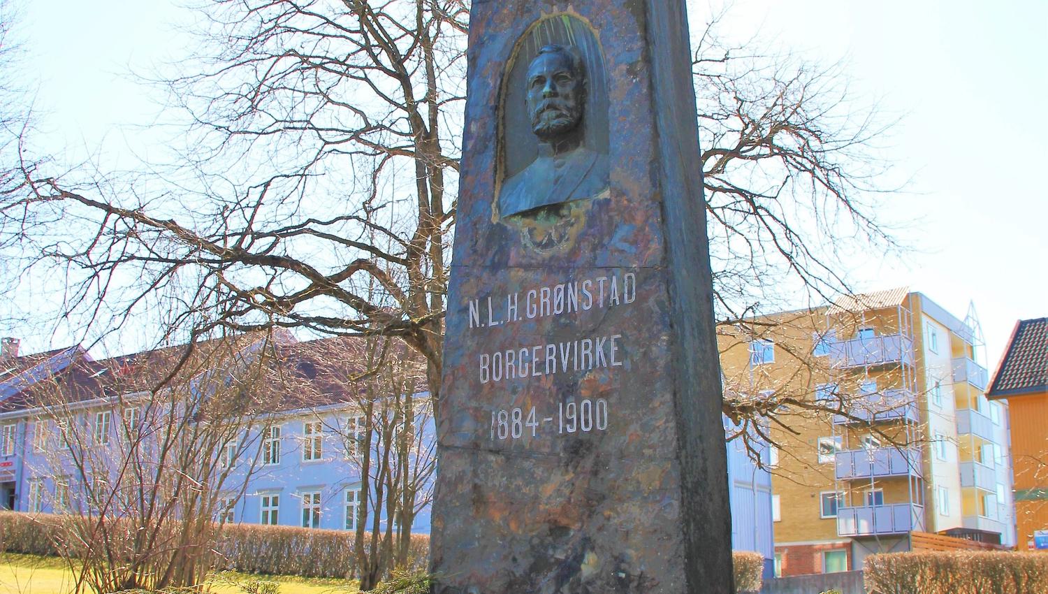 Høy steinmonument med inngravert tekst og nisje, omgitt av lave busker og trær ved boligblokker i sollys..