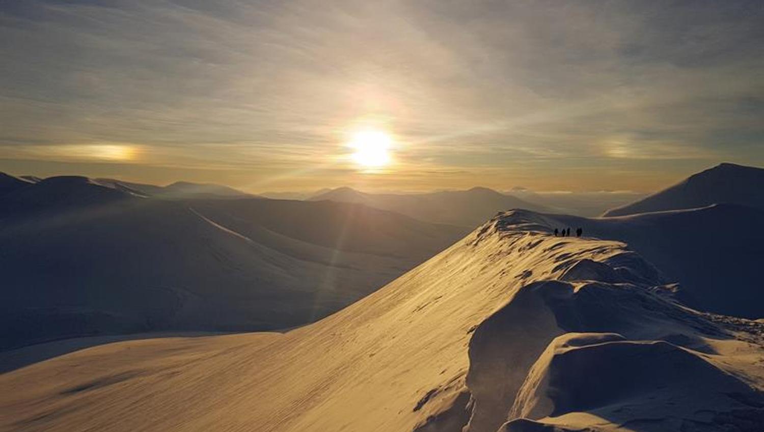 Sola som skinner på toppen av et snødekt fjell med nydelige fjell i bakgrunnen