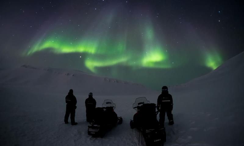 Three people standing next to two snowmobiles wathcing the northern lights on the sky