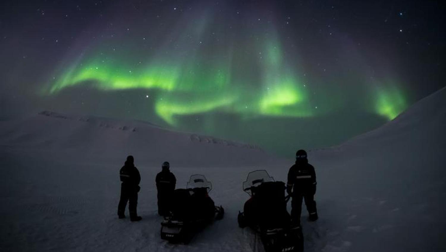 Three people standing next to two snowmobiles wathcing the northern lights on the sky
