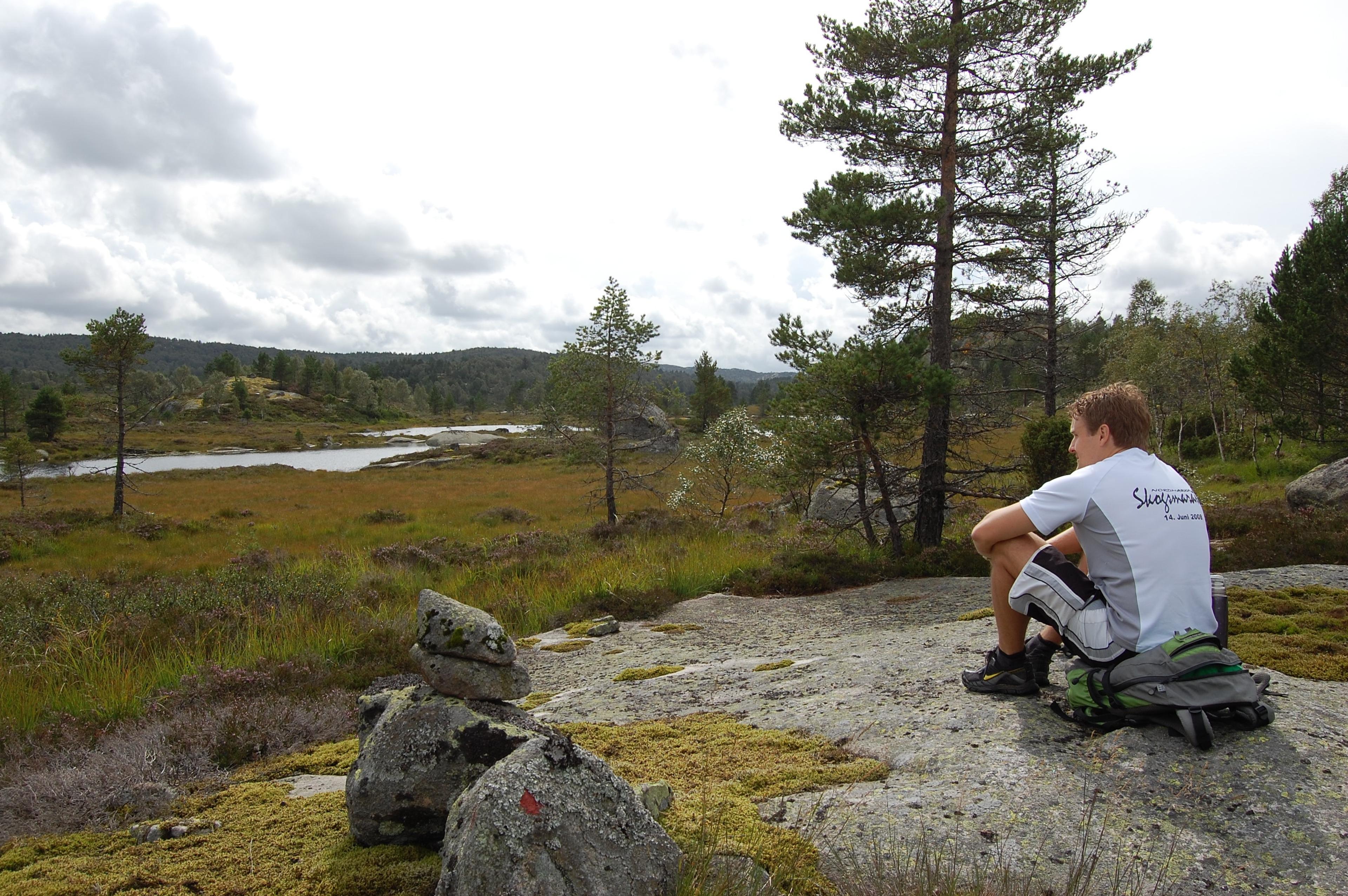 A man sitting on his backpack, looking out over the landscape with a water pond.